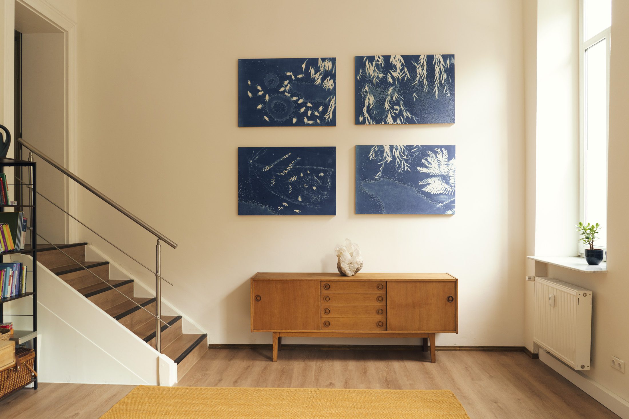 Blaue Pflanzenbilder über einem Holzsideboard im hellen Praxisraum. Links ist eine Treppe zu sehen.